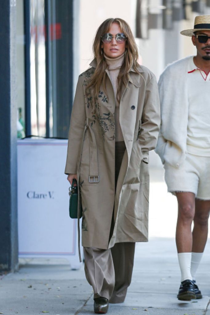 Jennifer Lopez walking down the street in a trench coat, sunglasses and matching trousers.