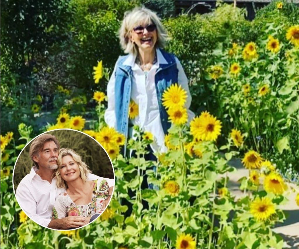 Olivia Newton John smiling in a white shirt and denim vest in a field of sunflowers