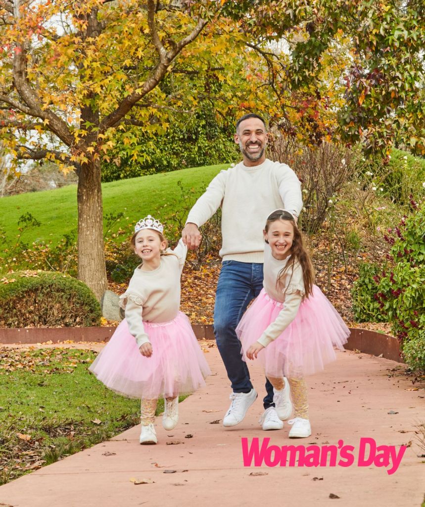 Adam and his two girls, smiling in a park, his girls holding his hands